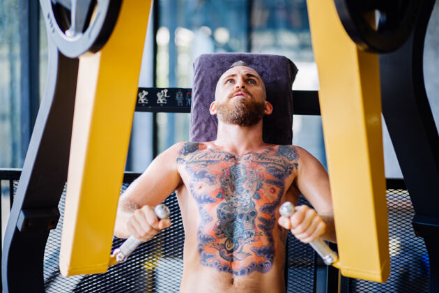 活跃在靠近窗户的健身房里 纹身的大胡子男人在胸部按压机上锻炼的肖像能量运动运动