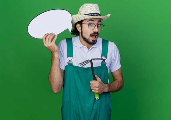 是年轻的留着胡须的园丁 穿着连体衣 戴着帽子 手里拿着床垫和一张空白的讲话气泡牌 惊讶不已绿色园丁帽子