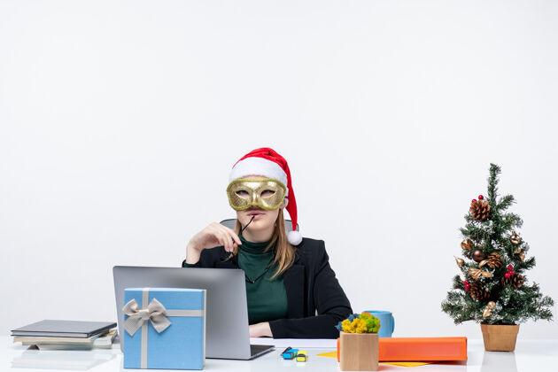 年轻人圣诞节的心情 自信的年轻女子戴着圣诞老人的帽子 戴着眼镜 戴着面具 坐在白色背景的桌子旁桌子办公室年轻女人