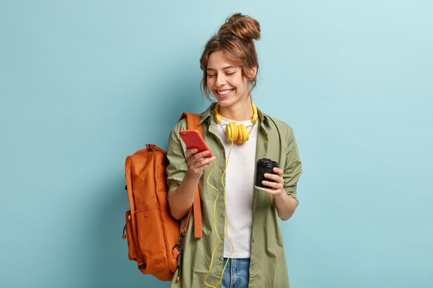 时髦人士穿着休闲服的积极年轻女子的照片 在手机上下载多媒体文件 脖子上戴着耳机正面智能手机乐观