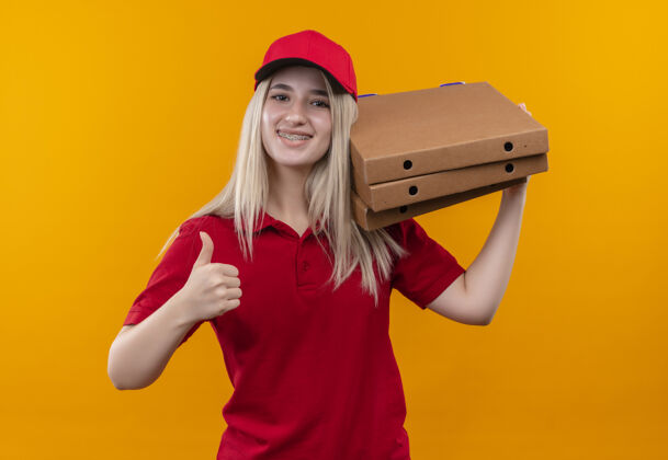 帽子微笑着送货的年轻女孩穿着红色t恤 戴着帽子 戴着牙套 肩上扛着披萨盒 大拇指竖立在孤立的橙色背景上女孩支架向上