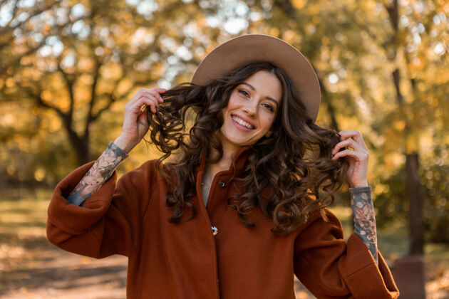 秋天画中有魅力的时尚微笑女子留着长长的卷发漫步在公园里穿着温暖的棕色外套秋日时尚 街头风格戴着帽子休闲优雅黄色
