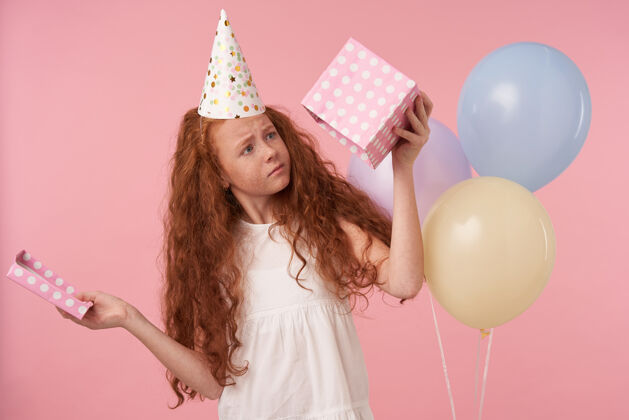 空气红头发卷发的女孩戴着生日帽庆祝节日的水平镜头 看着空荡荡的礼盒 失望地站在粉红色的工作室里 手里拿着彩色的气球女学生女性节日