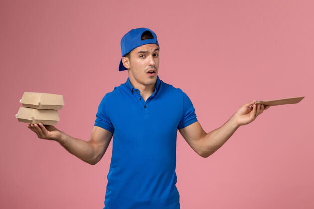 食物正面图：身穿蓝色制服披肩的年轻男性快递员拿着小快递食品包 在浅粉色的墙上放着记事本持有正面男人
