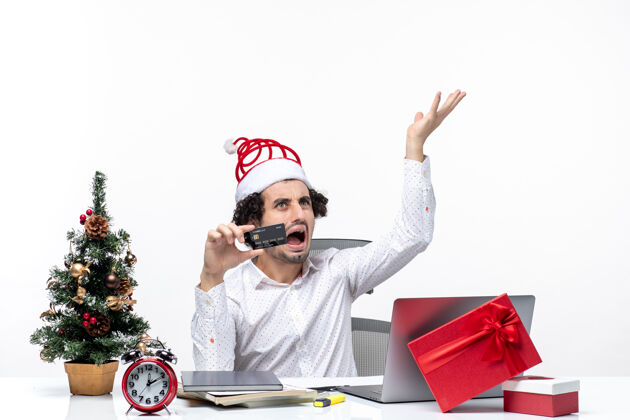 人在白色背景的办公室里 戴着圣诞老人的帽子 手里拿着银行卡的年轻疲惫愤怒的商务人士带着节日的喜庆心情圣诞老人节日圣诞节