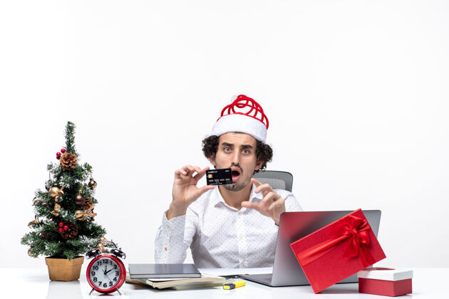 装置在白色背景的办公室里 戴着圣诞老人帽 拿着银行卡的商务人士感到震惊 节日气氛喜庆男性成人圣诞老人