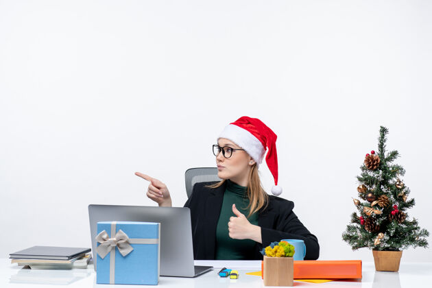圣诞节一位自信的商务女士 戴着圣诞老人帽 坐在一张桌子旁 桌上放着圣诞树和一份礼物 做着“ok”的手势 在白色背景下指着办公室右侧的东西一边笔记本电脑圣诞老人