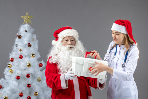 第一圣诞老人和女医生的前视图 女医生在灰色的墙上拿着他的急救箱服装女性圣诞老人