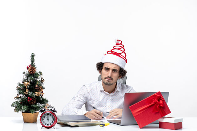 笔记本电脑专注的年轻商人 戴着有趣的圣诞老人帽子 在白色背景的办公室里检查庆祝圣诞节的便条微笑圣诞老人帽子