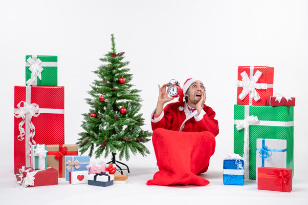 圣诞老人情绪激动的圣诞老人惊喜地看着上面坐在地上的钟 旁边的礼物和装饰圣诞树上的白色背景盒子礼物礼物