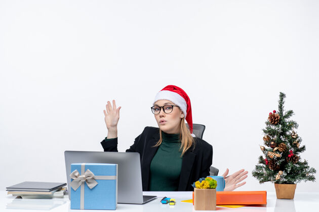 圣诞节一位严肃的年轻女士 戴着圣诞老人帽 坐在一张桌子旁 桌子上有一棵圣诞树和一份白色背景的礼物工作圣诞老人工作