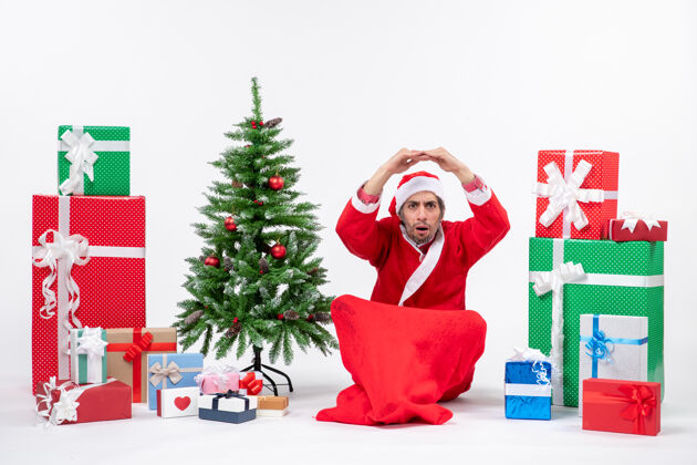 男人震惊的年轻人庆祝新年或圣诞节假期坐在地上附近的礼物和装饰圣诞树地坐震惊