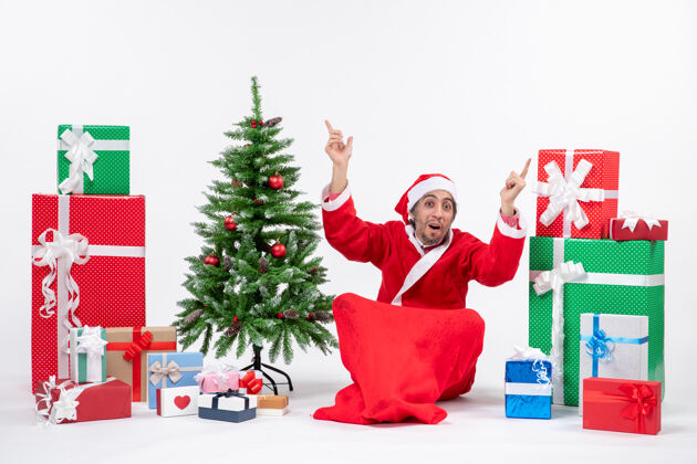 年轻人惊讶的年轻人装扮成圣诞老人 拿着礼物和装饰好的圣诞树坐在地上 在白色背景上指着两边的东西礼物东西圣诞