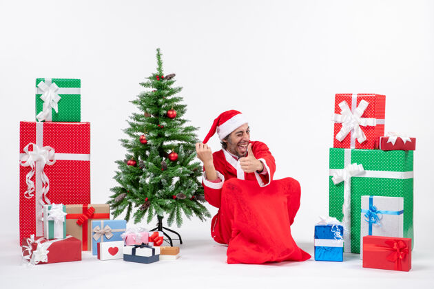 年轻人笑容可掬的年轻人打扮成圣诞老人 拿着礼物和装饰好的圣诞树坐在地上 在白色背景上做着ok的手势礼物圣诞树圣诞