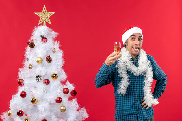 年圣诞心情与情绪激动的年轻人戴着圣诞老人的帽子 举起一杯葡萄酒欢呼自己圣诞树附近红色新年前夜圣诞