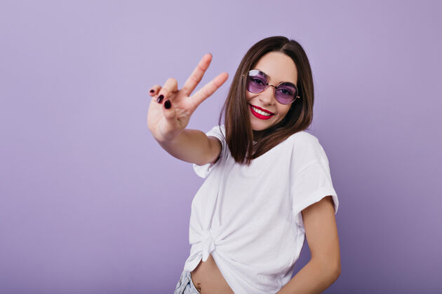 服装自信的欧洲女孩戴着圆眼镜摆出和平的姿势 微笑着优雅的白人妇女站在室内的照片紫色享受年轻