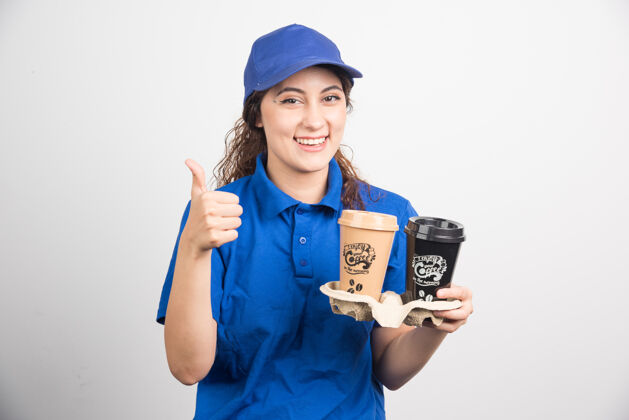 制服身着蓝色制服的女士举起大拇指 拿着两杯咖啡 背景是白色的高质量的照片帽子手女