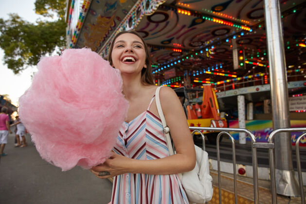 公园快乐迷人的年轻黑发女士 穿过游乐园 穿着夏装 手里拿着棉花糖 一边看一边开心地笑着女人长心情
