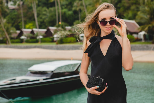 摆姿势奢华性感迷人的女人穿着黑色礼服在豪华度假酒店的码头上摆造型 戴着墨镜 暑假 热带海滩魅力海滩游艇