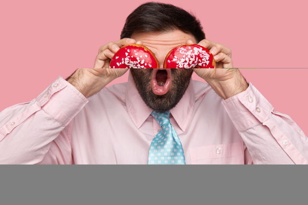 持有大胡子男人的特写镜头让红色的甜甜圈停留在眼睛上 让嘴巴张开可怕男性摆姿势