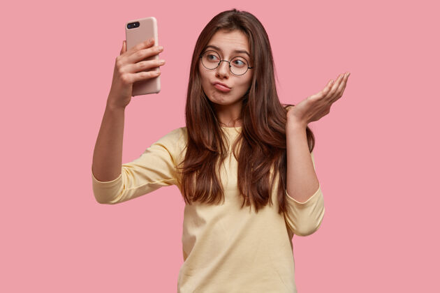 自拍漂亮犹豫不决的白人年轻女子形象以智能手机冷漠相看 自拍或打视频电话眼镜电子困惑