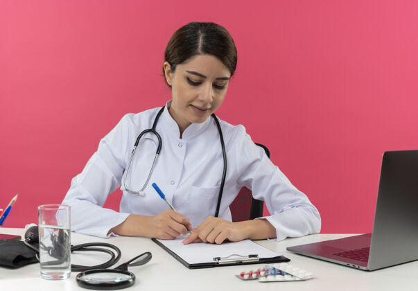 工作高兴的年轻女医生穿着医用长袍带听诊器坐在办公桌上用医用工具在剪贴板上写些东西医疗工具长袍