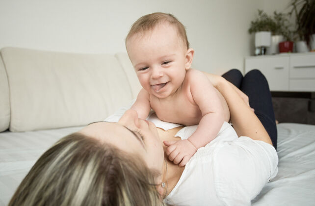 欢乐年轻微笑的母亲躺在床上和她6个月大的儿子玩耍的画像婴儿照顾女人