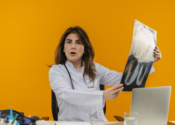 X光惊讶的中年女医生穿着医用长袍 带听诊器 坐在办公桌旁 用笔记本电脑和医疗工具 拿着x光片 背景是橙色 有复印空间工具穿着笔记本电脑