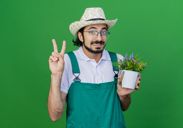 胡须年轻的留着胡须的园丁 穿着连体衣 戴着帽子 手里拿着一盆盆植物 上面写着“v”字看年轻园丁