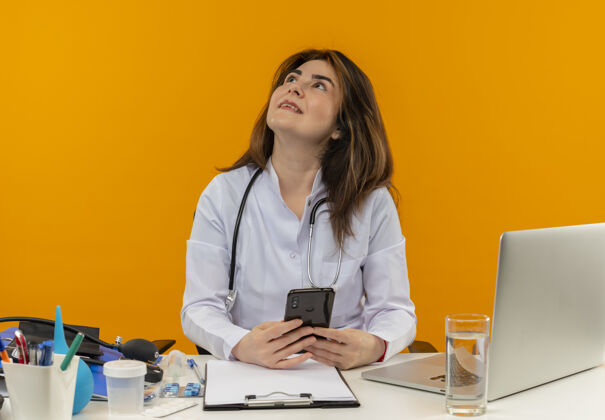 工具看着微笑的中年女医生 穿着医用长袍 手持听诊器 坐在办公桌旁 拿着笔记本电脑 拿着医疗工具 拿着电话 站在隔离的橙色墙上 还有复印空间看微笑电话
