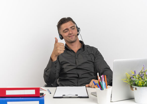 耳机年轻自信的金发上班族戴着耳机坐在办公桌旁 拿着办公工具 竖起大拇指看着笔记本电脑工人年轻看