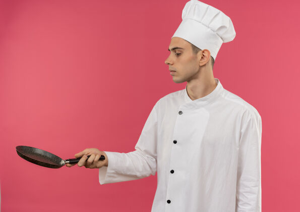 平底锅站在侧面年轻的男厨师穿着厨师制服拿着和看着煎锅与复制空间厨师男煎着