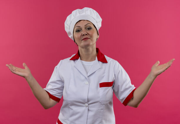 制服自信的中年女厨师身着厨师制服 在孤立的粉色墙上摊开双手蔓延厨师手