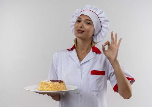 手势高兴的年轻厨师女穿着厨师制服 拿着蛋糕在盘子上 在隔离的白色墙壁上显示okey手势与复制空间好的年轻穿着