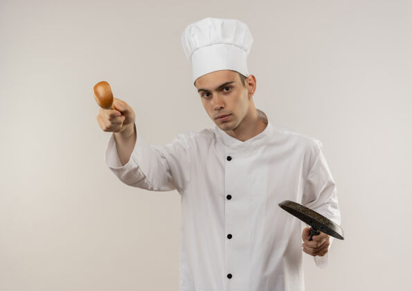 厨师自信的年轻男厨师穿着厨师制服 手拿煎锅 手拿勺子站在孤立的白色墙壁上厨师制服勺子