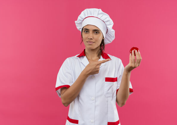 分年轻的女厨师穿着厨师制服 拿着西红柿点在隔离的粉红色墙上 留着复印空间厨师穿厨师