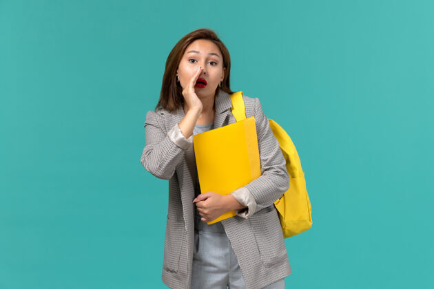 Female身穿灰色夹克 背着黄色背包 拿着文件在浅蓝色墙上低语的女学生的正面照片文件教训女学生