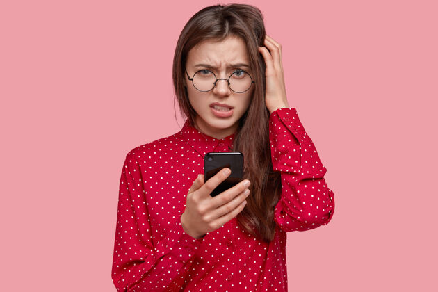 手机不高兴的黑发女人的照片困惑地看着 拿着手机 思考着留言的内容 穿着优雅的衣服消极手机抓挠
