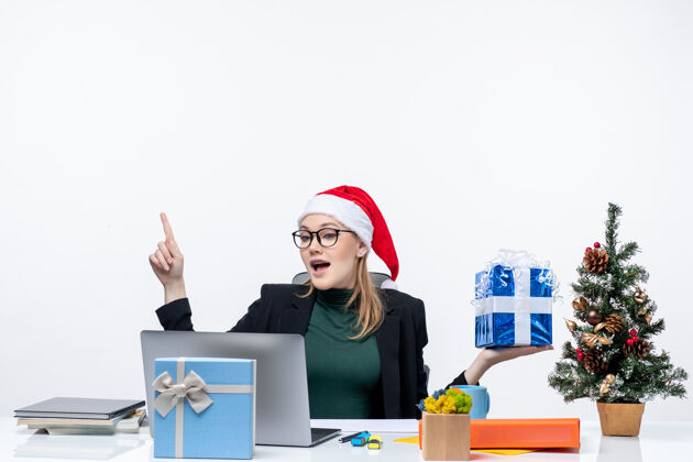 笔记本电脑圣诞气氛积极兴奋有趣的年轻女子戴着圣诞老人的帽子 戴着眼镜坐在一张桌子上 显示礼物指着上面的白色背景帽子眼镜圣诞老人