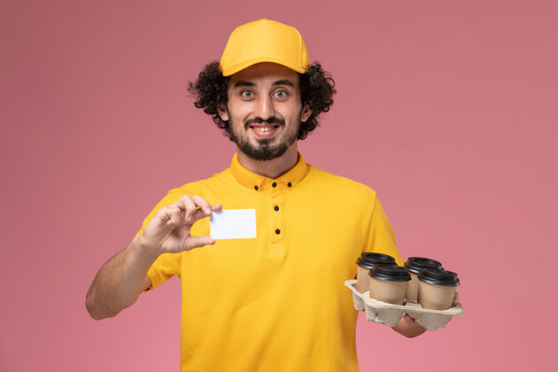 男快递正面图：身穿黄色制服的男信使手拿着棕色的咖啡杯和白色卡片放在粉红色的墙上专业制服杯子