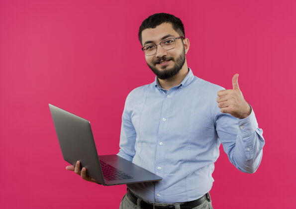 显示戴眼镜 穿蓝衬衫 留着胡子的年轻人拿着笔记本电脑微笑着竖起大拇指拿着向上衬衫