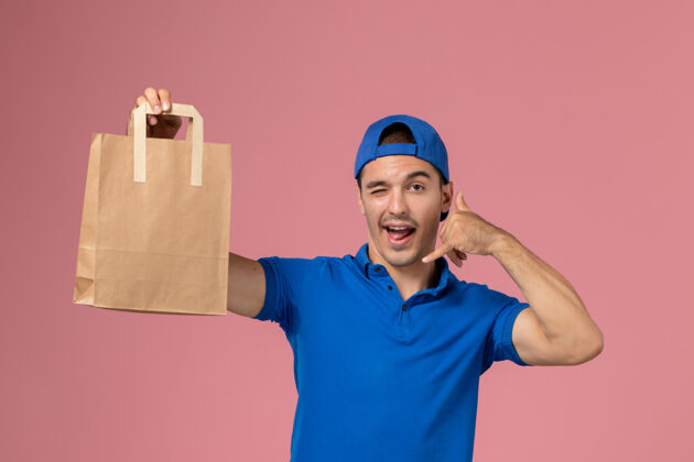 送货正面图：身穿蓝色制服和斗篷的年轻男性快递员 手上拿着快递纸包裹 放在粉红色的墙上工人粉色男性