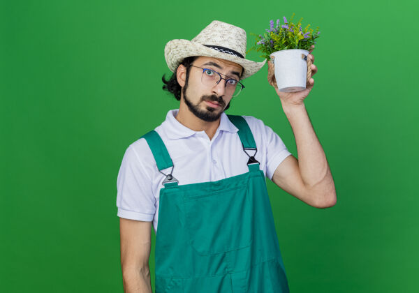 盆栽年轻的留着胡子的园丁 穿着连体衣 戴着帽子 头上拿着盆栽植物 困惑不解年轻看园丁