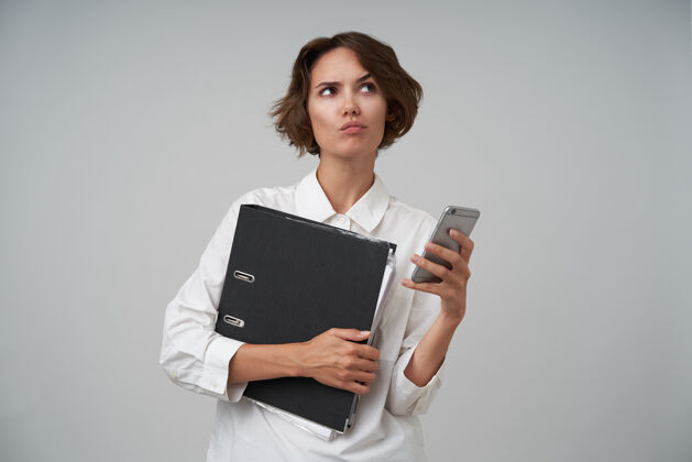 可爱一幅沉思的年轻黑发女性的肖像 穿着白衬衫 拿着文件和她的智能手机在办公室工作 若有所思地看着一边 扬起眉毛 站着抱着手机棕色