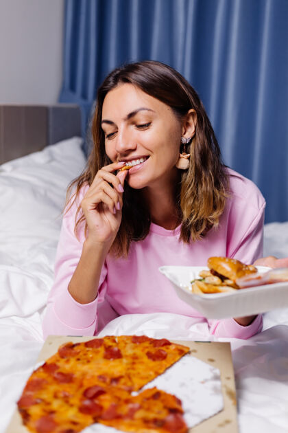 年轻女人东快餐从床上交货在卧室在家里的女性享受脂肪食品比萨饼和汉堡渴望碳水化合物睡衣服务送货