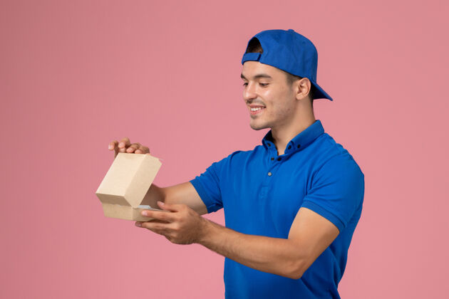 制服正面图：身穿蓝色制服披肩的年轻男性快递员在粉红色的墙上拿着小包裹正面视野快递