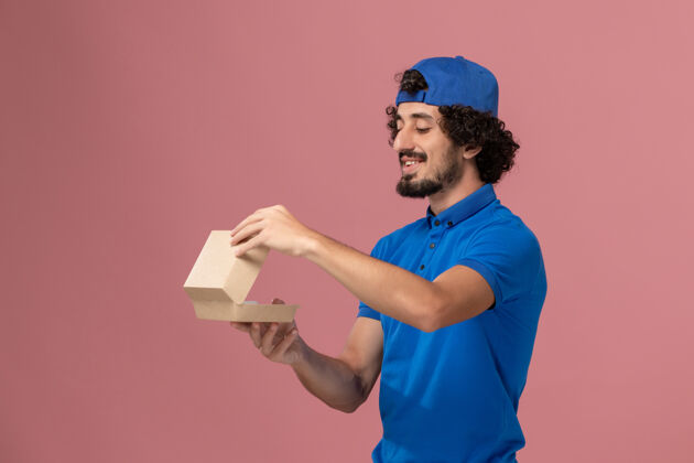 食物正面图：身穿蓝色制服的男性快递员 披肩上拿着一个小小的快递食品包 在粉红色的墙上打开它服务人手持