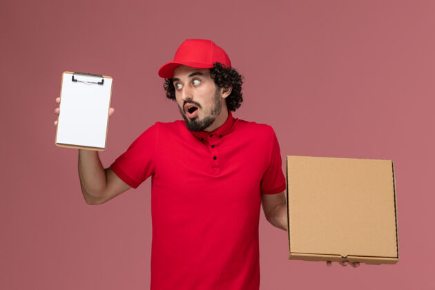 男性快递正面图：身穿红色衬衫和披风的男性快递员在粉色墙上拿着快递食品盒和记事本制服食物盒子