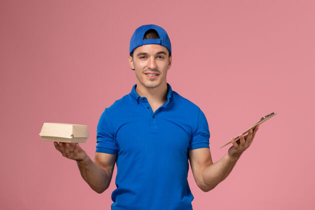 正面正面图身穿蓝色制服披肩的年轻男性快递员手持小快递食品包和笔记本在浅粉色的墙上食物快餐男人