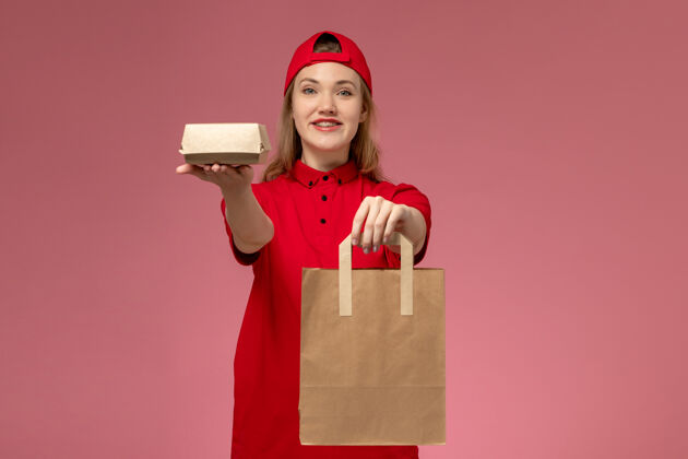 人正面图：身穿红色制服 披风的年轻女快递员在粉色墙上拿着快递食品包食物快递红色